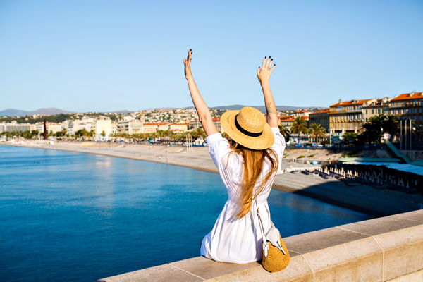 Na zdjęciu turystka w białej sukience i słomkowym kapeluszu nad brzegiem morza z uniesionymi w górę dłońmi